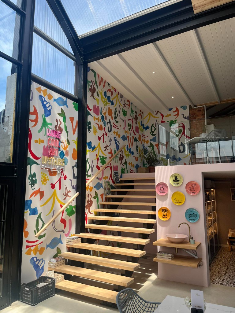 Abstract colourful wallpaper installation with neon signage reading More Cool Art Upstairs beside a staircase